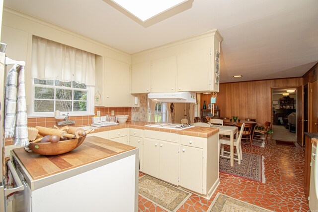 kitchen featuring white stovetop, tasteful backsplash, wooden walls, a peninsula, and under cabinet range hood