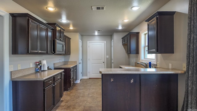 kitchen with light tile patterned floors, visible vents, stainless steel microwave, a peninsula, and dark brown cabinets