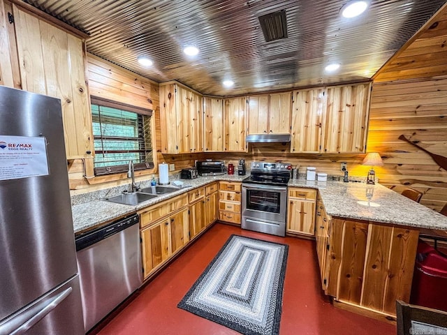 kitchen with wooden walls, appliances with stainless steel finishes, light stone counters, sink, and kitchen peninsula