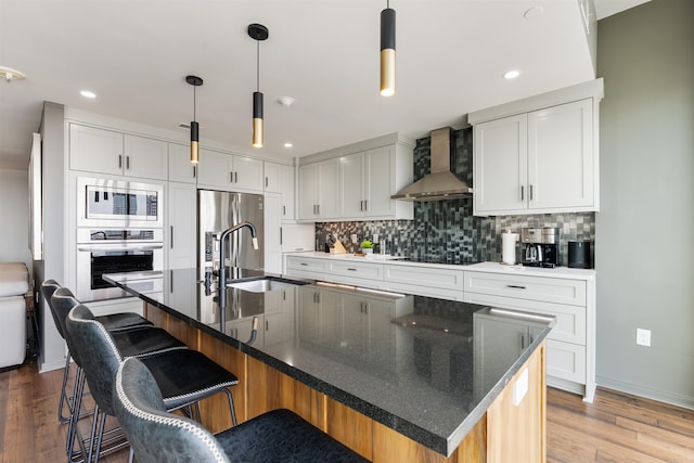 kitchen with stainless steel appliances, wall chimney exhaust hood, an island with sink, and white cabinetry