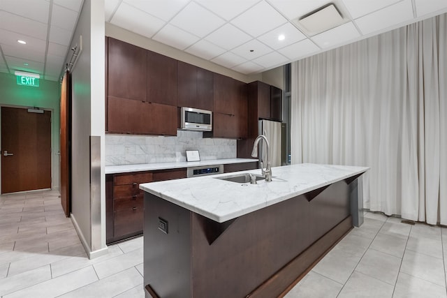 kitchen with light tile patterned floors, stainless steel appliances, sink, a drop ceiling, and a kitchen island with sink