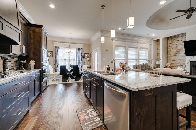 kitchen featuring an island with sink, sink, a stone fireplace, stainless steel appliances, and ceiling fan with notable chandelier