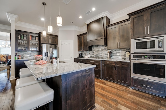 kitchen featuring premium range hood, an island with sink, hanging light fixtures, appliances with stainless steel finishes, and dark hardwood / wood-style floors