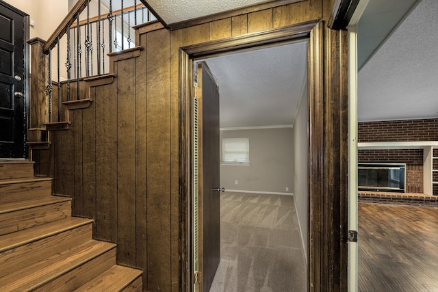 stairs featuring a brick fireplace, carpet floors, a textured ceiling, crown molding, and wooden walls