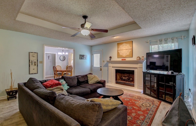 living room featuring ceiling fan with notable chandelier, light hardwood / wood-style floors, a tray ceiling, and a brick fireplace