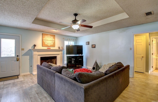 living room featuring light hardwood / wood-style floors, a brick fireplace, a raised ceiling, a textured ceiling, and ceiling fan