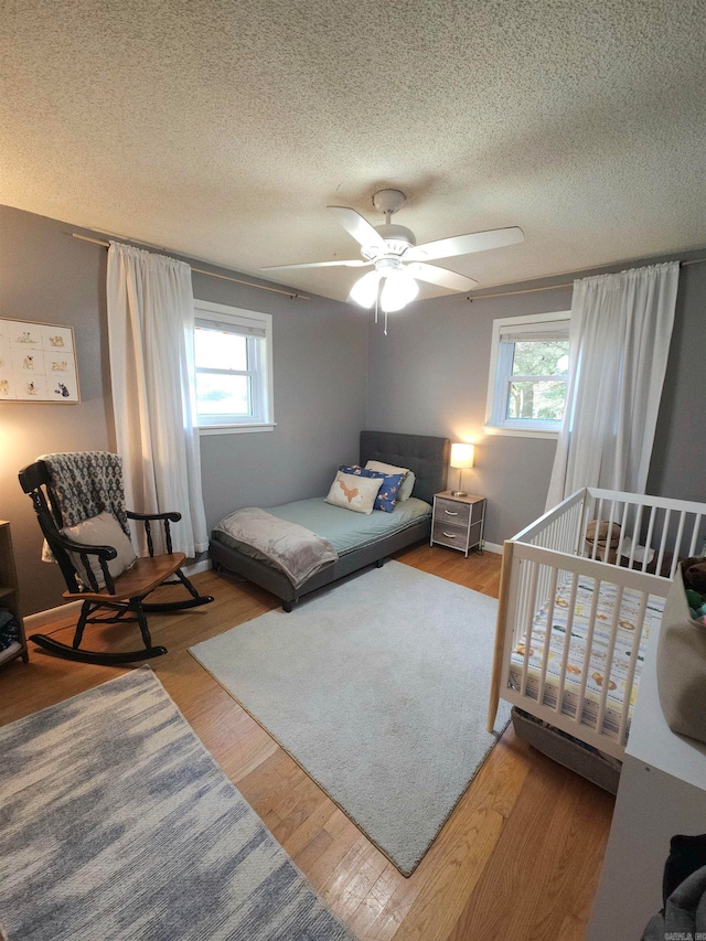 bedroom with a textured ceiling, ceiling fan, light hardwood / wood-style flooring, and a nursery area