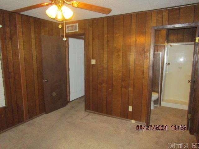 unfurnished bedroom featuring light carpet, wooden walls, ensuite bath, and ceiling fan