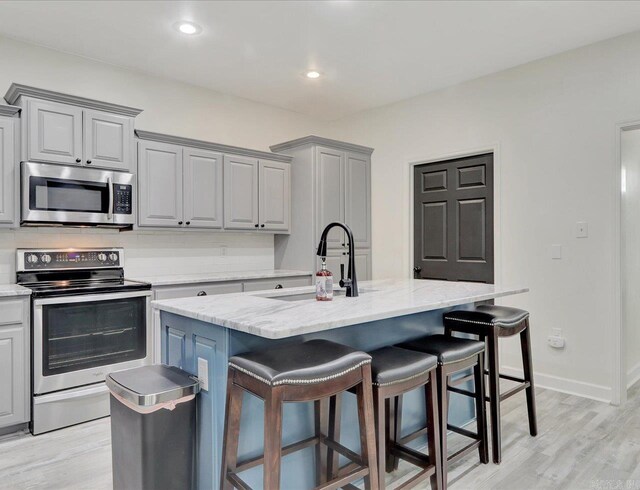 kitchen with appliances with stainless steel finishes, gray cabinetry, an island with sink, light wood-type flooring, and sink