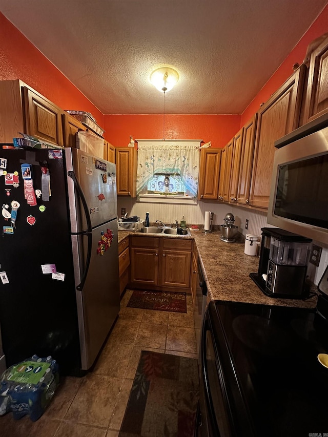 kitchen featuring appliances with stainless steel finishes, brown cabinetry, a sink, a textured ceiling, and dark tile patterned floors