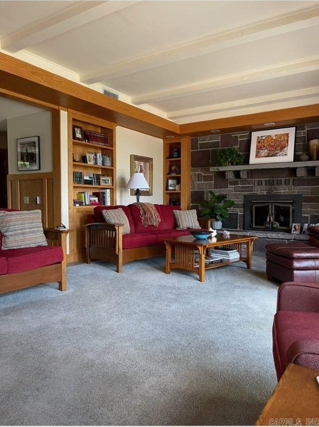 living room with a stone fireplace, carpet floors, built in shelves, and beamed ceiling