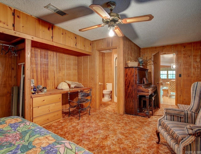 bedroom with ceiling fan, a textured ceiling, wooden walls, light colored carpet, and ensuite bath