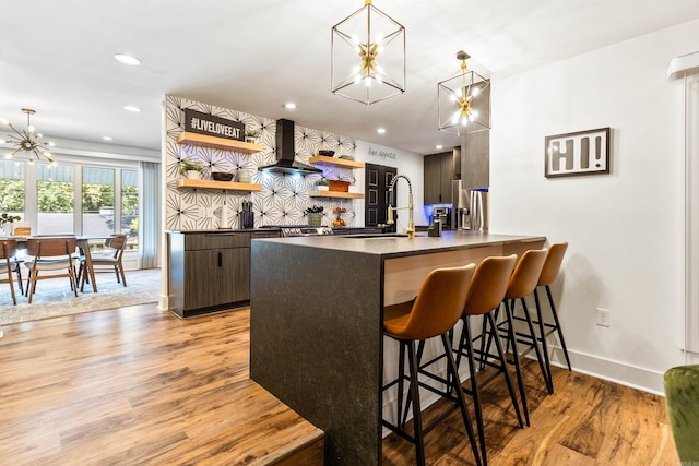 bar featuring hanging light fixtures, sink, dark brown cabinets, light hardwood / wood-style flooring, and wall chimney range hood