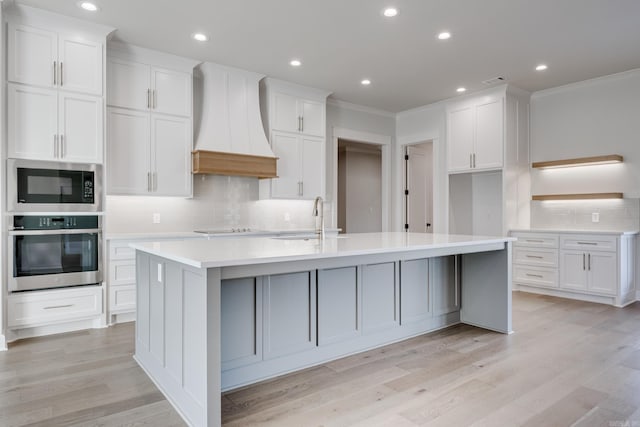 kitchen with premium range hood, appliances with stainless steel finishes, white cabinets, and an island with sink