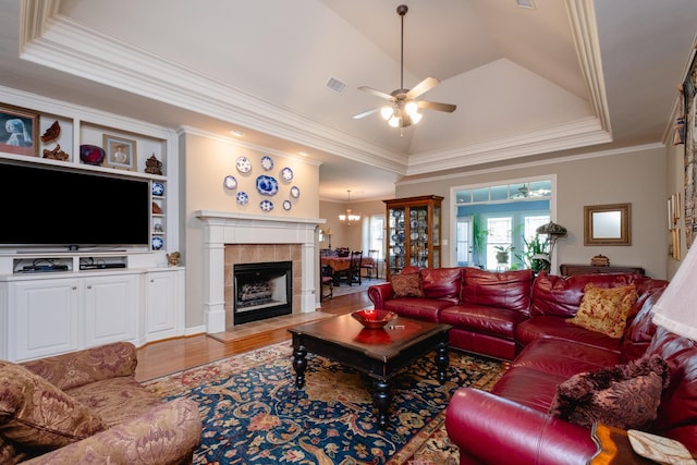 living room featuring a tiled fireplace, ceiling fan with notable chandelier, light hardwood / wood-style flooring, a raised ceiling, and ornamental molding