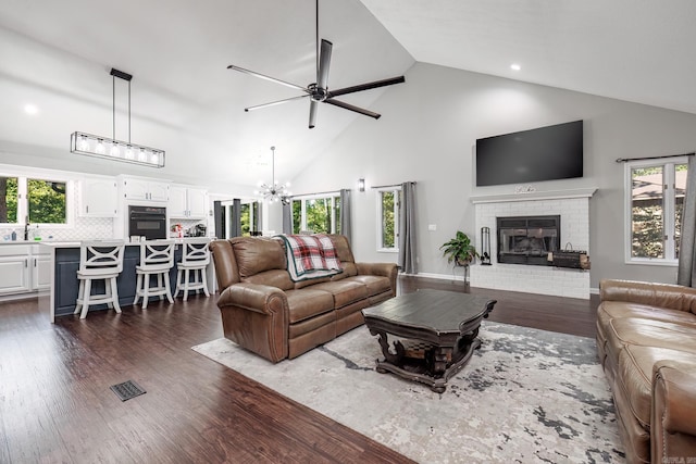 living room featuring a brick fireplace, high vaulted ceiling, sink, dark hardwood / wood-style flooring, and ceiling fan with notable chandelier