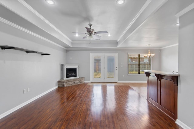 unfurnished living room with a brick fireplace, ceiling fan with notable chandelier, a tray ceiling, dark hardwood / wood-style floors, and ornamental molding