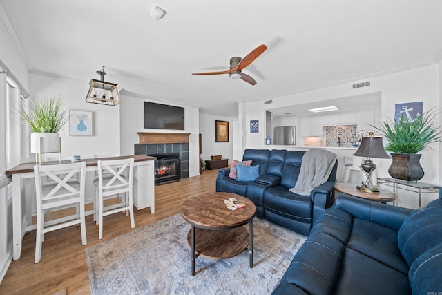 living room featuring ceiling fan, a tiled fireplace, light hardwood / wood-style floors, and crown molding