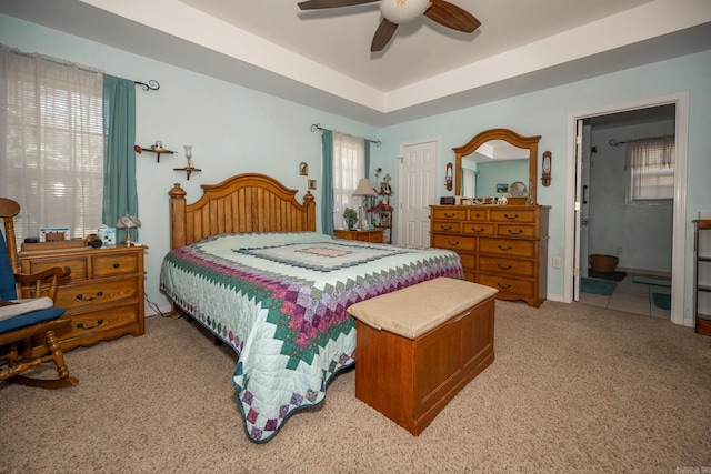 carpeted bedroom featuring ceiling fan, a tray ceiling, multiple windows, and ensuite bath