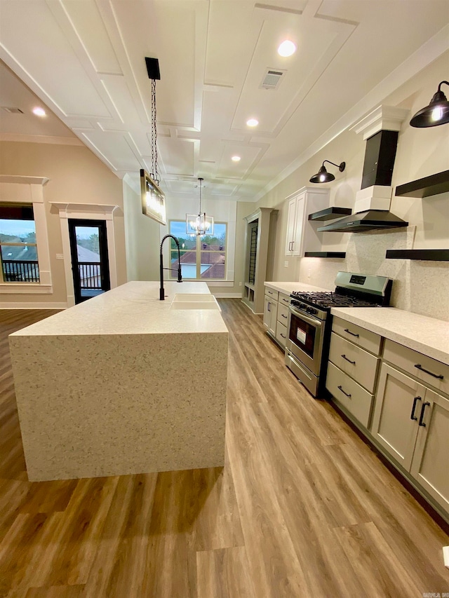 kitchen featuring gas stove, wall chimney range hood, hanging light fixtures, an island with sink, and light hardwood / wood-style floors