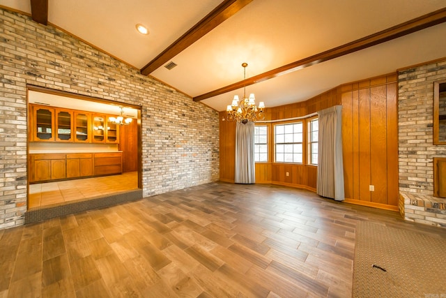 unfurnished living room with an inviting chandelier, wood-type flooring, wood walls, lofted ceiling with beams, and brick wall