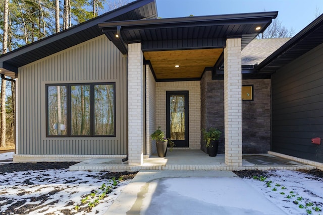 snow covered property entrance with a porch, brick siding, a shingled roof, and board and batten siding