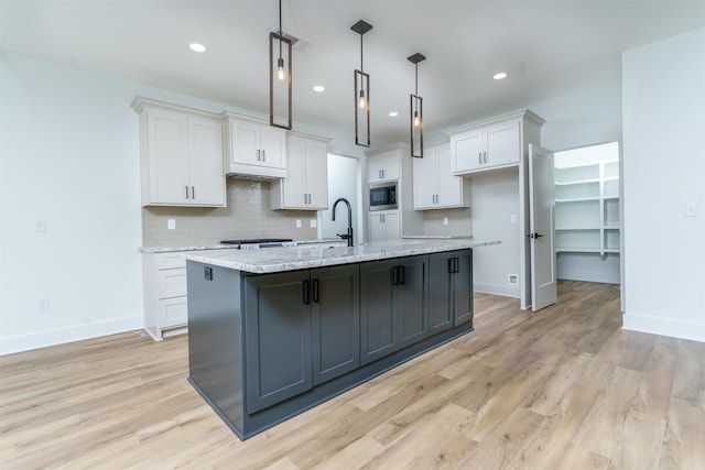 kitchen featuring white cabinets, hanging light fixtures, light stone countertops, a kitchen island with sink, and built in microwave