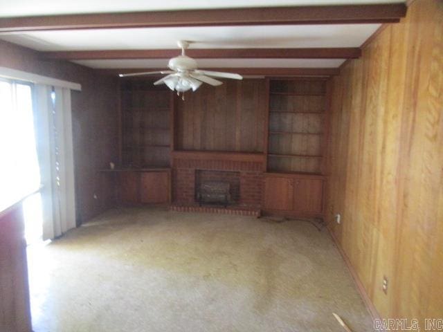 unfurnished living room featuring ceiling fan, beamed ceiling, a brick fireplace, and wooden walls