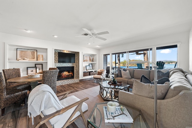 living room with built in shelves, dark wood-type flooring, a tiled fireplace, and ceiling fan