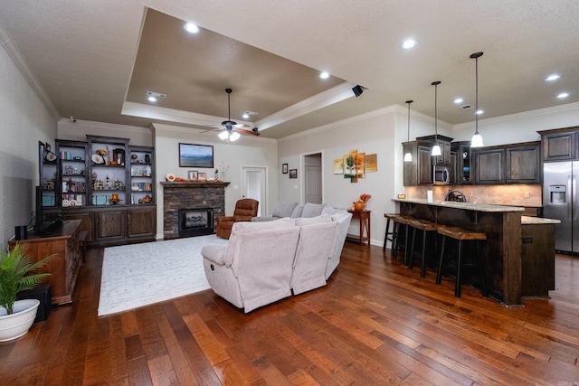 living room featuring a raised ceiling, ceiling fan, dark hardwood / wood-style floors, a stone fireplace, and crown molding