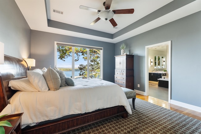 bedroom with hardwood / wood-style flooring, ensuite bath, a raised ceiling, a water view, and ceiling fan