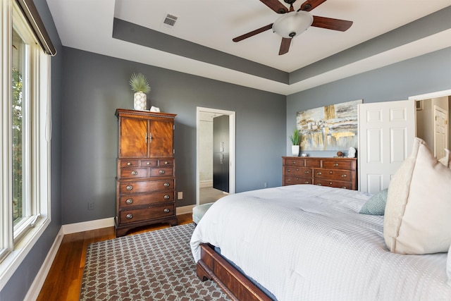 bedroom with a tray ceiling, dark wood-type flooring, ensuite bath, and ceiling fan