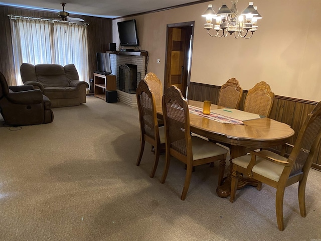 dining area with carpet flooring, a brick fireplace, ceiling fan with notable chandelier, wooden walls, and crown molding