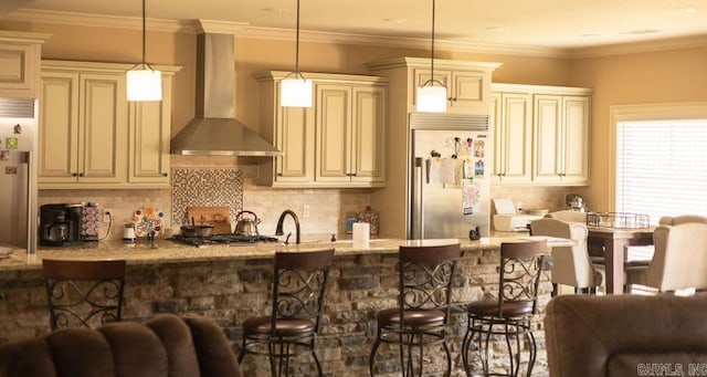 kitchen featuring cream cabinetry, wall chimney exhaust hood, hanging light fixtures, light stone countertops, and appliances with stainless steel finishes