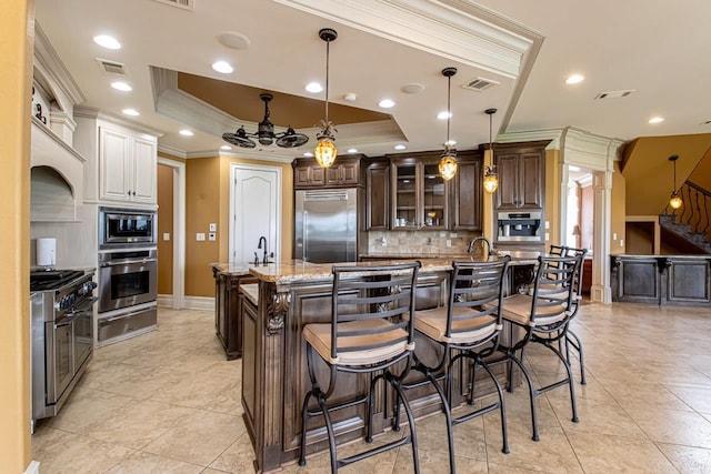 kitchen with a spacious island, a raised ceiling, hanging light fixtures, built in appliances, and light stone counters