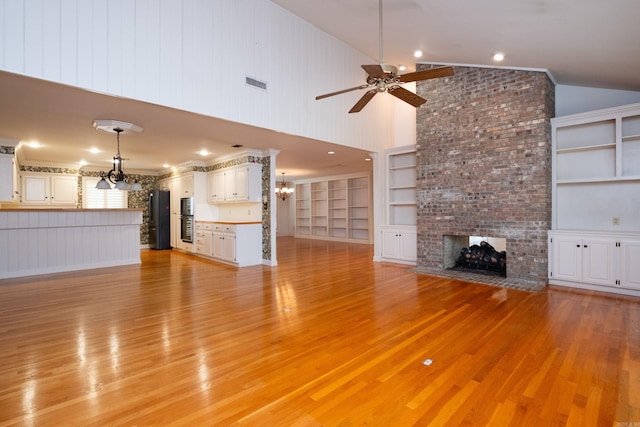 unfurnished living room with light wood-type flooring, ceiling fan with notable chandelier, a brick fireplace, high vaulted ceiling, and built in shelves