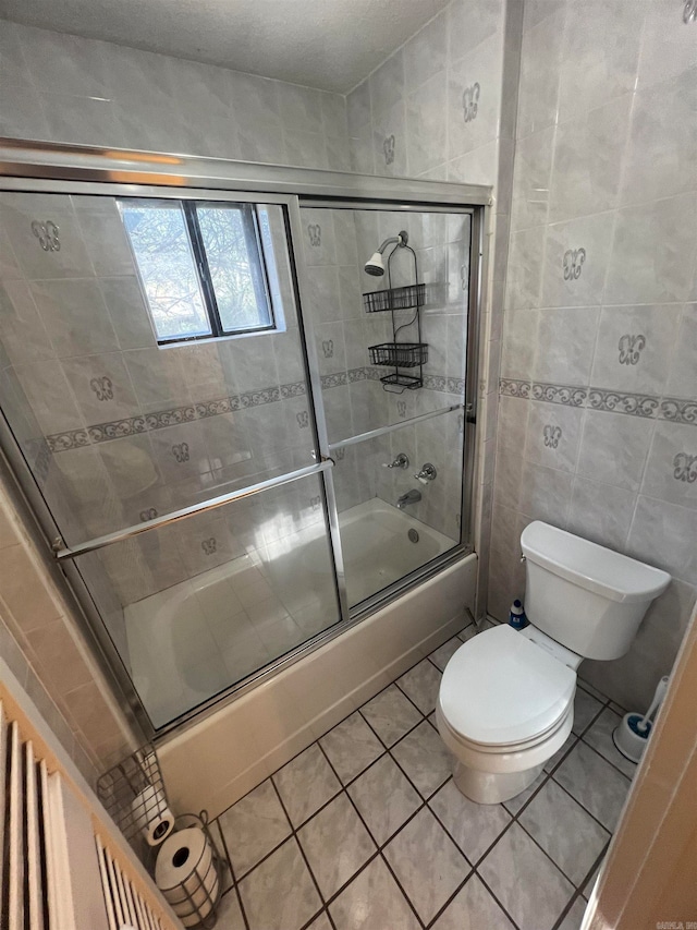 bathroom with tile walls, tile patterned floors, bath / shower combo with glass door, and toilet