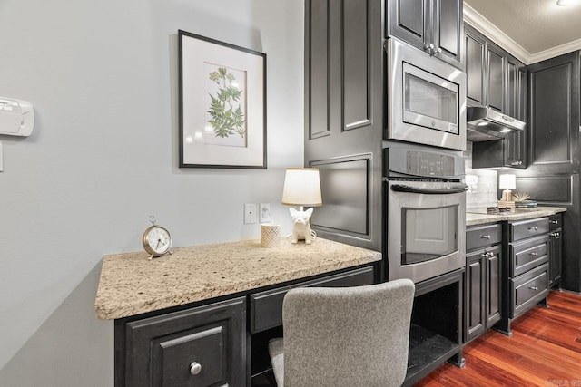 kitchen with built in desk, dark hardwood / wood-style flooring, appliances with stainless steel finishes, crown molding, and light stone counters