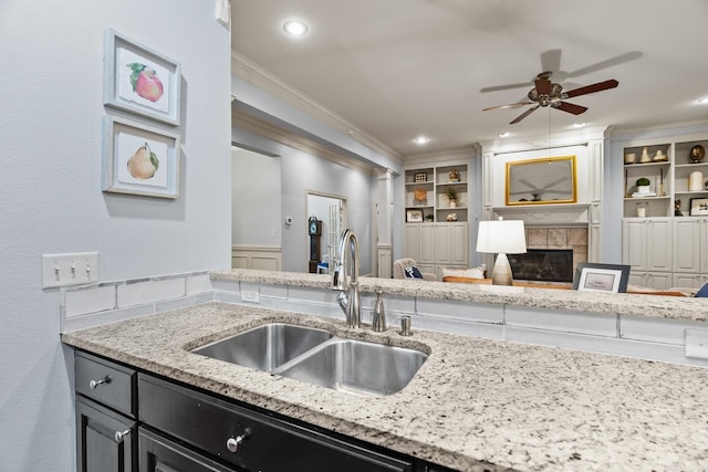 kitchen with sink, a tiled fireplace, ceiling fan, light stone counters, and ornamental molding