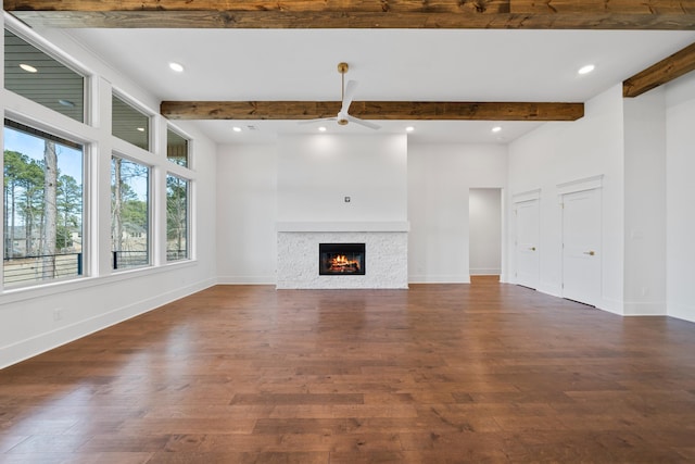 unfurnished living room featuring ceiling fan, dark hardwood / wood-style floors, beamed ceiling, and a stone fireplace