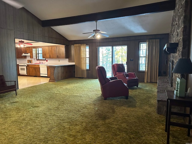 carpeted living room featuring wood walls, vaulted ceiling with beams, ceiling fan, and a stone fireplace