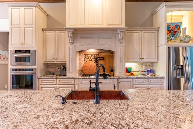 kitchen with backsplash, appliances with stainless steel finishes, light stone countertops, cream cabinetry, and sink