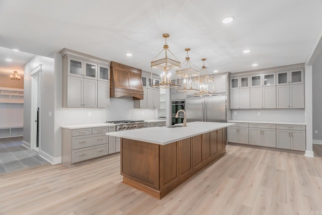 kitchen with light hardwood / wood-style floors, an island with sink, custom range hood, appliances with stainless steel finishes, and decorative light fixtures