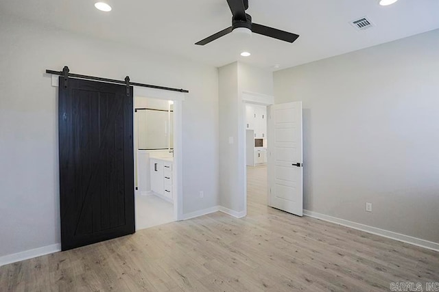 unfurnished bedroom featuring ensuite bath, light hardwood / wood-style floors, a barn door, and ceiling fan