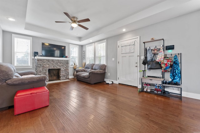 living room with ceiling fan, wood-type flooring, a stone fireplace, and a tray ceiling