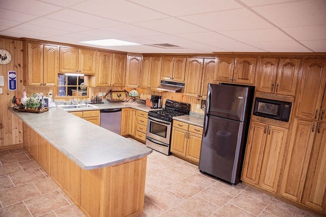 kitchen with a drop ceiling, stainless steel appliances, kitchen peninsula, wood walls, and sink