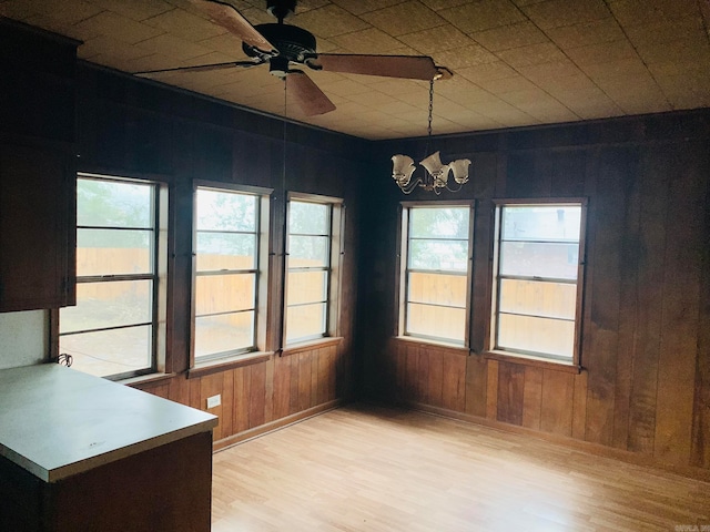 unfurnished dining area with ceiling fan with notable chandelier, light hardwood / wood-style flooring, and wooden walls