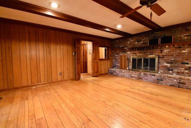 unfurnished living room featuring a brick fireplace, light hardwood / wood-style floors, wooden walls, ceiling fan, and beam ceiling