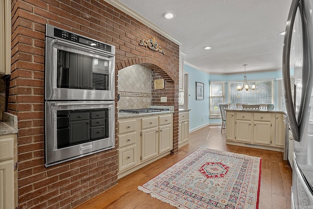 kitchen featuring stainless steel appliances, ornamental molding, decorative light fixtures, cream cabinetry, and light wood-type flooring