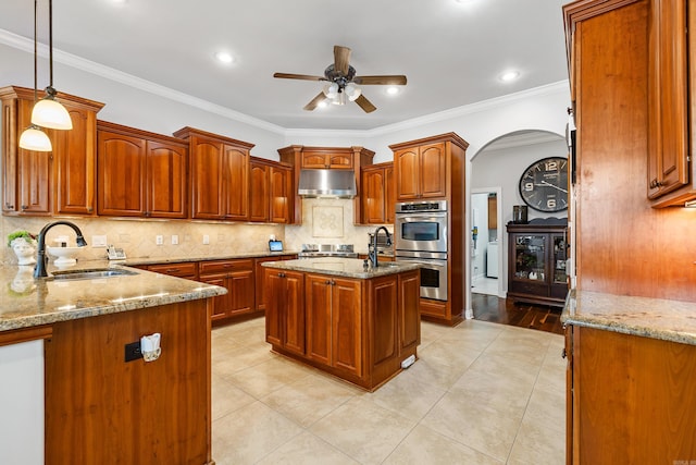 kitchen with double oven, a kitchen island with sink, crown molding, sink, and hanging light fixtures